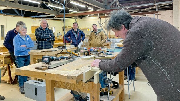 Students standing around a table looking at a man demonstrate a plane during a how to become a boat builder course