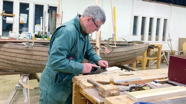 A man in green overall using plane on a piece of wood