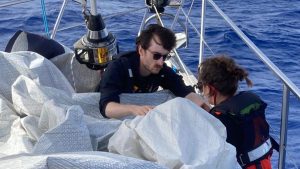 Two people wearing lifejackets making boat repairs while at sea, specifically sewing up a split sail