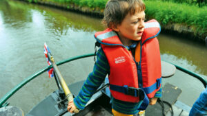 A child wearing a personal floatation device at the tiller of a boat. A lifejacket is essential inland gear for exploring the waterways