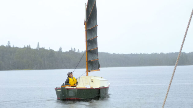 A boat with a junk rig being sailed on a lake by a man wearing a lifejacket