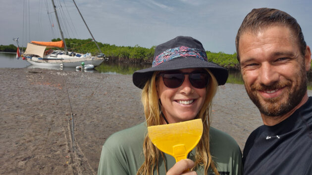 A couple holding a plastic scraper to clean the hull of their boat which is careening in a river