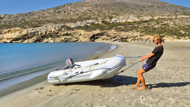 A man wearing a black tshirt and denim shorts pulling a PVC dinghy up a beach
