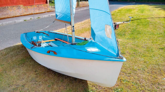 A blue and white cruising dinghy on grass with the sails up
