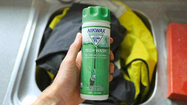 A hand holding a bottle of Nikwax Tech Wash above sailing gear ready for washing. Credit: Gill/gb.gillmarine.com