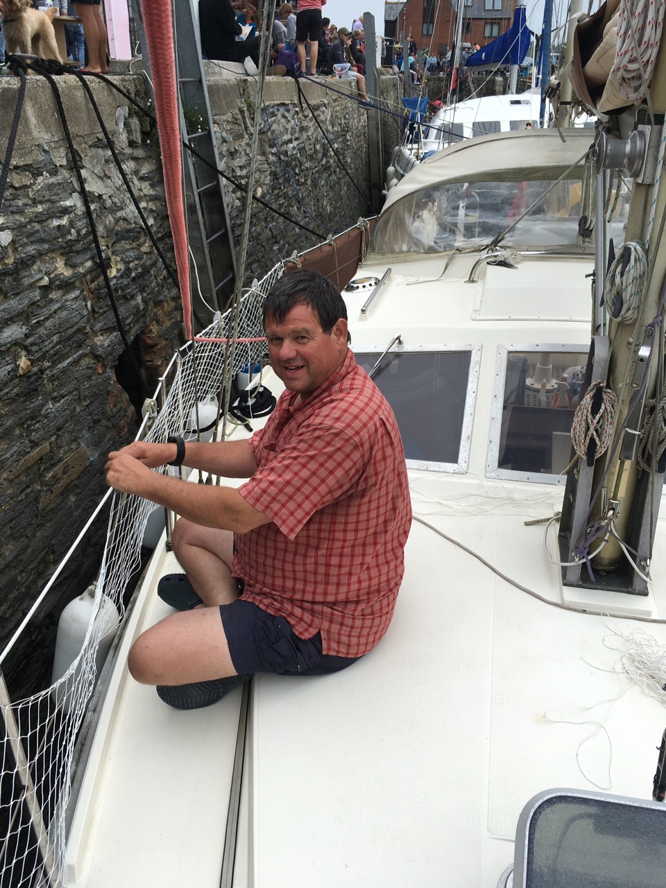 Jonty Peace in a red checked shirt and shorts sitting on the foredeck of his cat