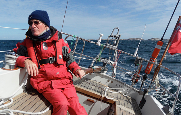 Dr Roger Chisholm dressed in red with a dark hat and sunglasses helming a boat