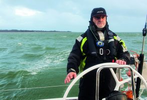 A skipper, dressed in a black cap and wet weather gear helming a yacht