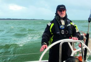 A skipper, dressed in a black cap and wet weather gear helming a yacht