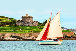 Small yacht with red and white sails passing St Mawes Castle on the Roseland peninsula in Cornwall UK