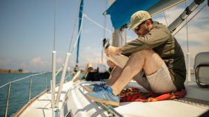 A man sitting on the deck of a yacht wearing a cap reading adventure books