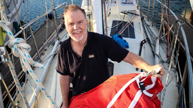 2018 Golden Globe Race skipper, Mark Sinclair wearing a black tshirt and holding an orange sail bag