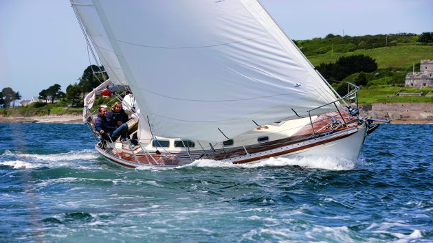 A Rustler 36 with white hull and sails heeling while sailing past the coastline