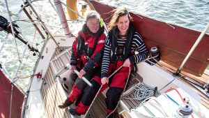 Two women wearing lifejacekts on a boat while it is heeled over. US Sailing is urging all sailors to wear one