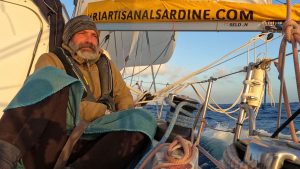 A sailor wearing a hat and a beard sitting on the deck of his boat sailing through crazy seas