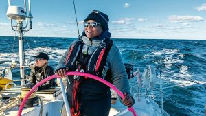 Lungi wears sunglasses, a hat, fleece, and life jacket. She is stood at the helm, steering. The weather is bright, with some clouds.