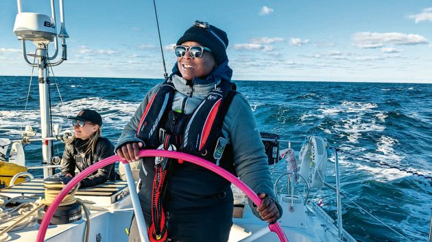 Lungi wears sunglasses, a hat, fleece, and life jacket. She is stood at the helm, steering. The weather is bright, with some clouds.