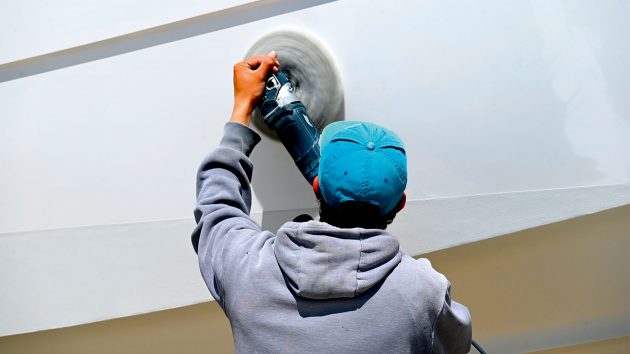 A man is holding a circular power tool on a pole up to the hull. He is polishing the hull.