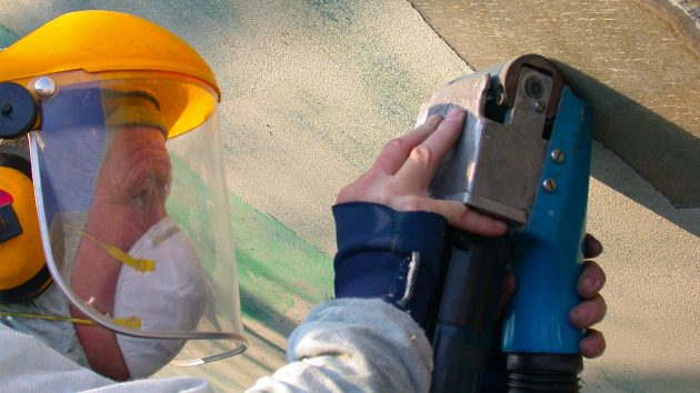 A man is wearing a protective visor, mask and hard hat as he uses a power tool against a hull.