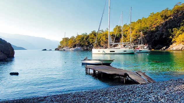 The yacht is moored in a secluded cove with trees behind and a rocky beach in front.