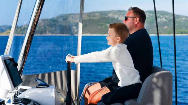 A child sat on someone's lap steering at the helm