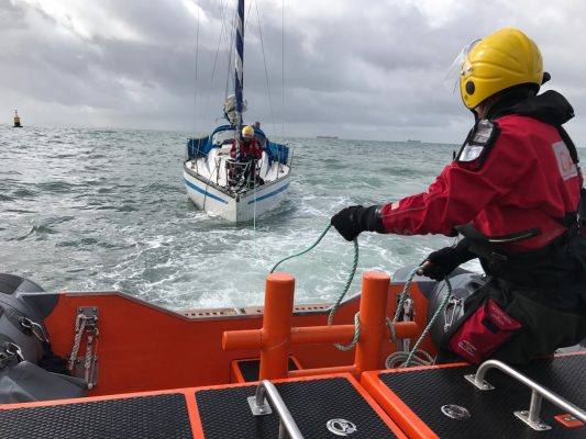 A yacht with propeller problems is towed by an orange lifeboat