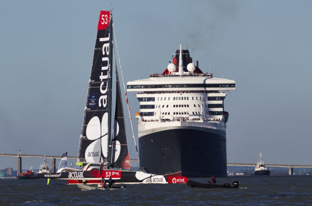 A maxi trimaran with red, black and white sails passes in front of an ocean liner