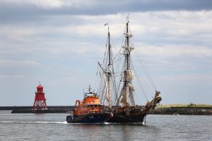 A masted sailing ship - Tres Hombres is towed by an orange lifeboat