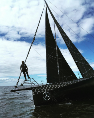 A man stands on the bow of a black IMOCA 60