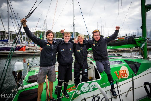 Four crew members on a green, orange and white yacht celebrate after finishing a race