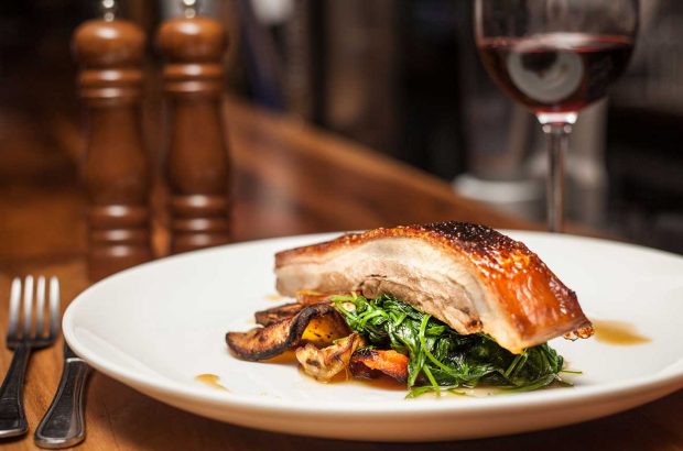 A glass of red wine with a dish of pork belly and vegetables.