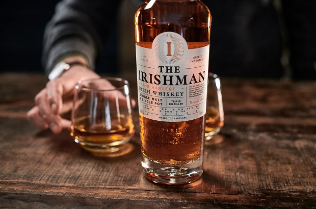 A hand holds a glass of whisky on a wooden bar next to a bottle of whisky