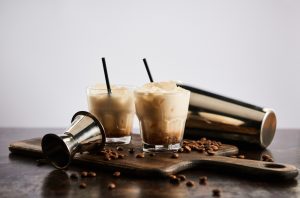 Two White Russian cocktails on a wooden board with coffee beans and a cocktail shaker