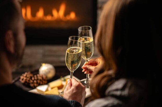 Two people with glasses of sparkling wine and cheese board in front of fire